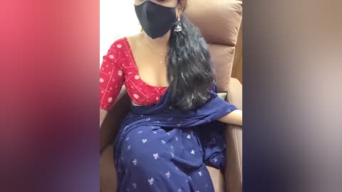 Video of a South Asian woman wearing a red top with white patterns, a black mask, and a blue sari, sitting in a tan recliner chair.