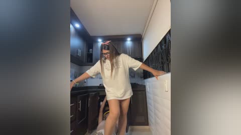 Video of a young woman with long hair, wearing a white oversized t-shirt and no pants, dancing energetically in a modern kitchen with dark cabinets and white countertops.