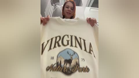 Video of a young woman with long, straight auburn hair, smiling, holding a white T-shirt with \"Virginia\" and a moose graphic, in a modern room with a white ceiling fan.