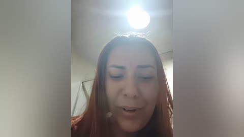 A video of a young woman with long, reddish-brown hair and fair skin, captured from the chest up, smiling with eyes closed, under a bright light.