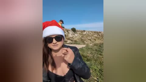 A video of a woman in a Santa hat and sunglasses, standing outdoors on a rocky hillside. She has a slender physique and is wearing a black top. In the background, a man in a yellow shirt stands further up the hill.