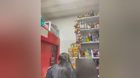 Video of a woman with long black hair, wearing a black shower cap, standing in a well-stocked convenience store with shelves full of snacks, drinks, and a red cooler.