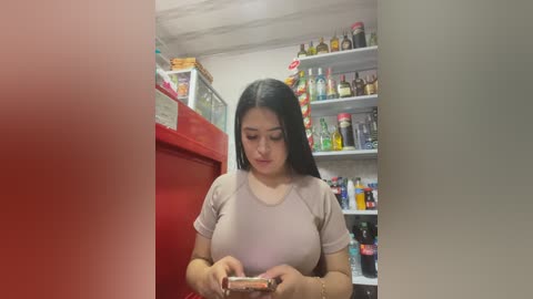 Video of a young woman with long black hair, medium skin tone, wearing a light pink t-shirt, looking at a jar in a cluttered, brightly-lit convenience store.