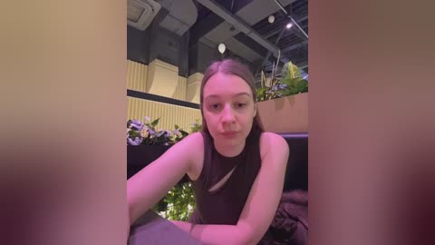 Video of a young Caucasian woman with light skin and long brown hair, wearing a sleeveless black dress, sitting in a modern, indoor setting with green plants, yellow walls, and industrial ceiling lights.