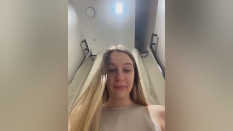 A video of a young Caucasian woman with long, straight blonde hair, wearing a light-colored, sleeveless top, standing in a narrow, dimly lit hallway with gray walls.