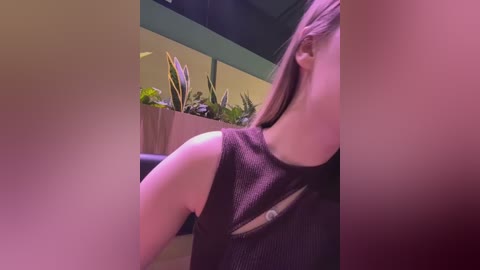 Video of a woman with light skin and long brown hair, wearing a black sleeveless dress, leaning against a wall with green plants in a potted planter.