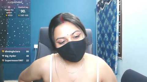 A video of a woman with dark hair and a black mask, wearing a white tank top, sitting in a chair in a blue room with a blue curtain and a weather forecast screen.