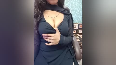 Video of a curvy woman with long, wavy dark hair, wearing a deep V-neck black dress, revealing ample cleavage. She stands in an office with a teal wallpaper and patterned cubicle divider.