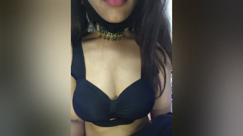 Video of a woman with medium skin tone wearing a black bra, a black choker with golden beads, and a black off-shoulder top. Her long, dark hair falls past her shoulders. The background is blurred and neutral-toned.