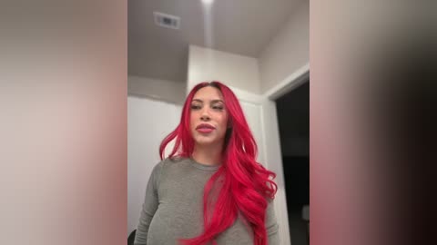 Video of a woman with long, vibrant red hair and fair skin, wearing a grey long-sleeve top, standing in a modern hallway with white walls and doors.