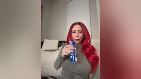Video of a woman with long, vibrant red hair, holding a blue can of Diet Coke, standing in a modern, minimalist living room with beige walls and a white couch.