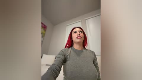 Video of a woman with long red hair, fair skin, and large breasts, wearing a grey long-sleeved top, standing in a modern, white-walled room with a floral painting in the background.