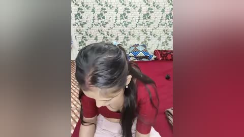 A video shows a young woman with long black hair in braids, wearing a red blouse and white skirt, kneeling on a red bedspread. The background features a green floral wallpaper and a wicker basket.