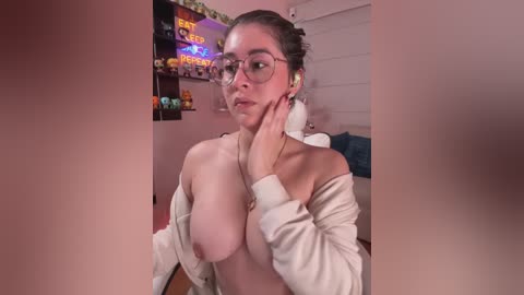 Video of a topless, fair-skinned woman with large breasts, wearing glasses, and a beige robe, in a dimly lit room with a neon sign and a bed.