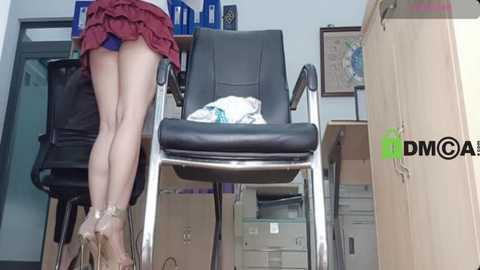 Video of a woman in a red mini-skirt, exposing her bare legs and buttocks, standing in an office. Behind her, a black leather office chair with clothes on it, bookshelves, and a DMMCA logo.