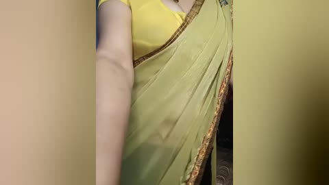 A close-up video of a woman's upper body in a green sari with gold embroidery, wearing a yellow blouse, captured from a slightly blurry angle, indoors.