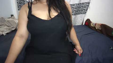 Video of a curvy woman with long dark hair, wearing a tight black tank top, sitting on a bed with blue sheets and patterned pillows, in a cozy, dimly lit bedroom.
