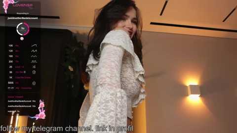 A video of a dark-haired woman in a sheer lace top, standing in a dimly lit room with a wall-mounted light fixture.