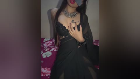 Video of a woman with long dark hair, wearing a black lace bra and sheer black dress, sitting on a floral-patterned bed. She has a silver choker and red lipstick, with a blindfold covering her eyes.