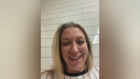 Video of a smiling Caucasian woman with long blonde hair, wearing a white shirt with vertical black stripes, in a tiled bathroom with white walls and a mirror.