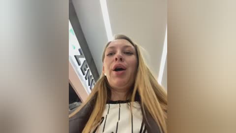 Video of a blonde woman with long hair, wearing a black jacket over a white blouse, mouth open in what appears to be a moment of surprise or excitement.