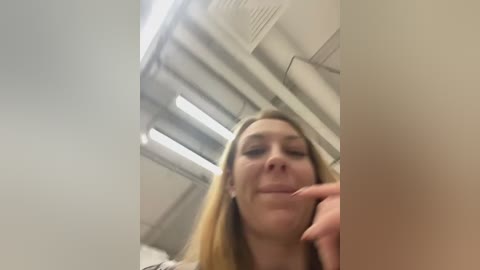 Video of a young Caucasian woman with long blonde hair, smiling, in an airplane overhead compartment. The background shows fluorescent lights, metal ceiling panels, and luggage.