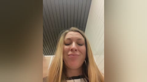 Video of a young Caucasian woman with long blonde hair, closed eyes, and a neutral expression. She stands in a dimly lit, modern hallway with beige walls and a dark, textured ceiling.