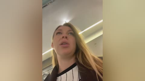 Video of a blonde woman with fair skin, wearing a black and white striped shirt, looking upwards in an indoor setting with fluorescent lights.
