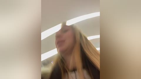 A blurred video of a young woman with long blonde hair, smiling, captured from a low angle, standing under a fluorescent-lit ceiling in a modern, clean setting.