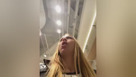 Video of a blonde woman with long hair, looking upwards, in a dimly lit, modern hallway with white walls and fluorescent lighting.
