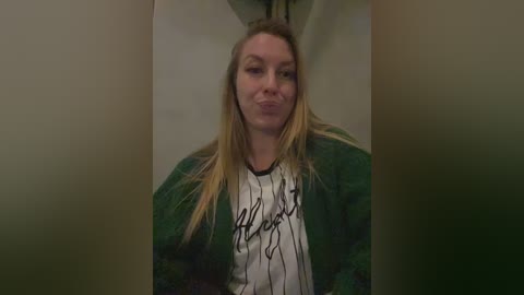 Video of a blonde woman with straight hair, wearing a green patterned hoodie and white T-shirt, making a kissy face, indoors with blurred background.
