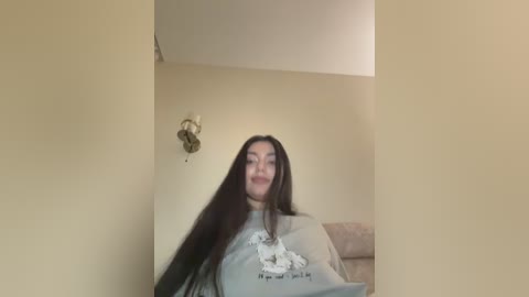 A video of a young woman with long, dark hair, wearing a grey sweatshirt, standing in a beige-walled room with a sconce light and a sofa in the background.