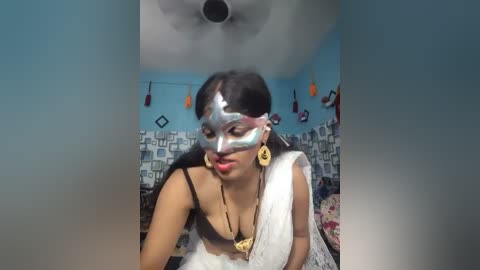 A video of a woman with dark skin, dressed in traditional Indian attire, wearing a silver mask and large gold earrings. She is indoors with blue walls, a ceiling fan, and patterned curtains.