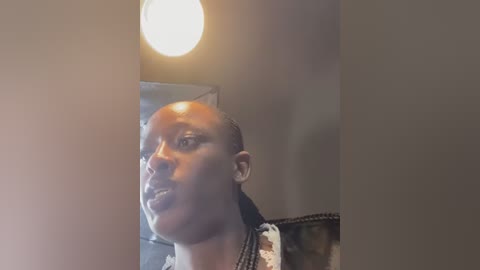 A video of a bald, light-skinned African woman with braided hair, wearing a black and white patterned dress, standing in a dimly lit room with a glowing light bulb overhead.