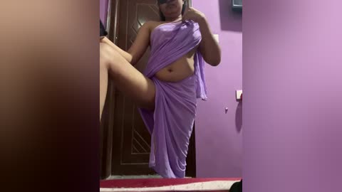 A video of a South Asian woman with medium skin tone, wearing a purple saree, lifting her leg in a mirror. Background features purple walls and a wooden door.