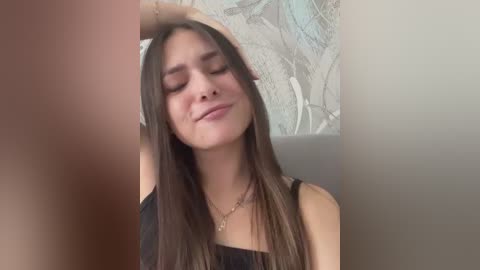 Video of a young woman with long, straight dark hair, light skin, and closed eyes, wearing a black top, standing in a room with a muted, abstract wall pattern.