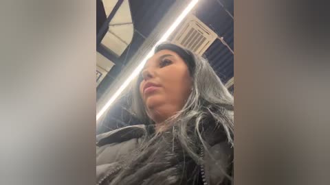 Video of a woman with long, silver hair wearing a grey puffer jacket, taken from a low angle inside a modern building with industrial lighting and exposed beams.
