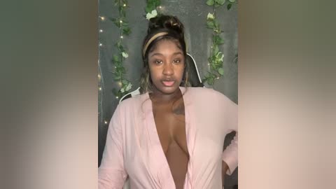 Video of a young Black woman with dark skin, medium-sized breasts, wearing a light pink button-down shirt with a deep V-neck, against a background of green ivy and fairy lights.
