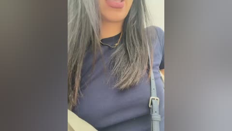 Video of a woman with long, straight, silver hair, wearing a dark blue T-shirt, and a grey crossbody bag. Her lips are slightly parted, hinting at a playful or surprised expression. The background is blurred.