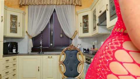 A video of a stylish kitchen featuring cream cabinets, a purple window with sheer white curtains, a black chair, and a woman in a red fishnet top.