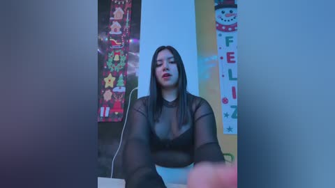 Video of a Latina woman with long black hair, wearing a sheer black top, seated in a room decorated with holiday-themed posters and lights, creating a festive, intimate atmosphere.
