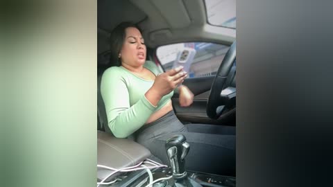 Media: Video of a woman in a car, wearing a green top and gray pants, with a cellphone in hand, looking irritated, with blurred background.