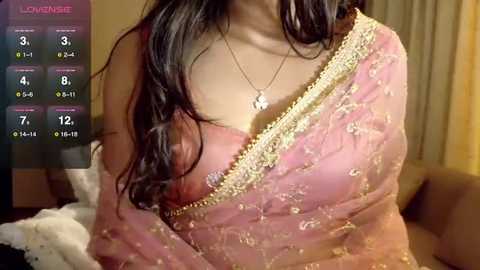 Video of a South Asian woman with long dark hair, wearing a pink saree with gold embroidery, in a cozy indoor setting.