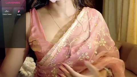 Video of a woman in a pink saree with gold embroidery, wearing a white mask, hands touching her chest, in a dimly lit room with beige curtains.