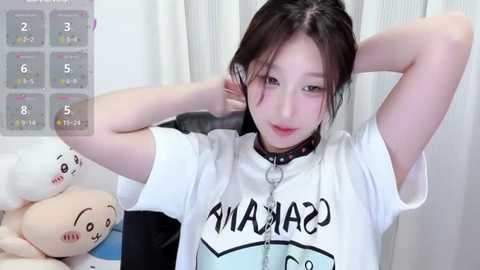 Video of an East Asian woman with fair skin and dark hair, wearing a white T-shirt with \"CARNIVAL\" and a choker. She's in a room with white curtains and stuffed animals.