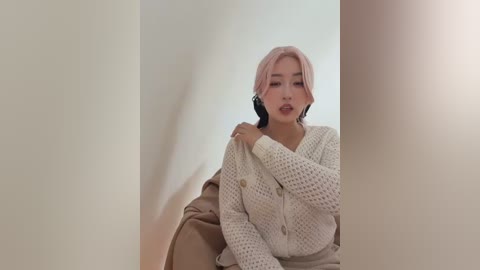 Video of an East Asian woman with light skin and pink hair, wearing a white knitted cardigan and beige jacket, seated against a plain white background, looking contemplative.