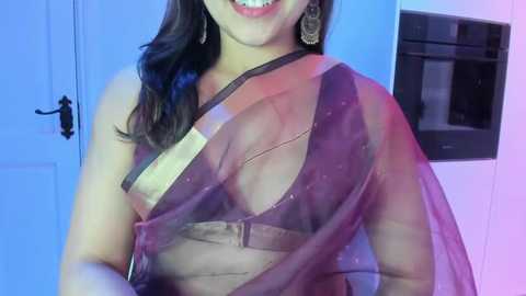 Video of a smiling South Asian woman with medium brown skin, wearing a sheer purple sari with a gold belt, large hoop earrings, and a gold necklace. Background shows a blue wall with a microwave and door.
