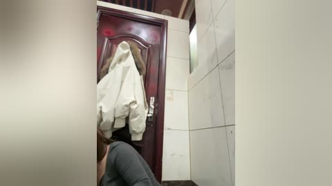 A video of a bathroom with a person in a gray shirt holding a white towel, leaning against a wooden door with a red knob, and white tiled walls.