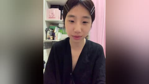 A video of an East Asian woman with light skin, wearing a black robe, hair in two black hair clips, standing in a cluttered bathroom with a pink shower curtain and shelves holding toiletries.