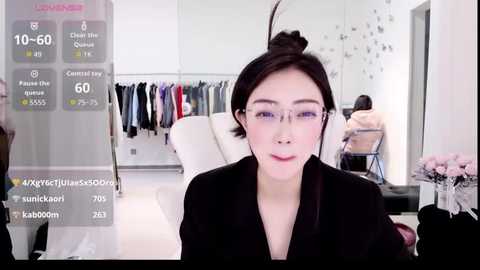 A video of an East Asian woman with fair skin, dark hair in a bun, and glasses, sitting in a modern, well-lit room. She wears a black outfit and is engaged in a virtual reality experience.